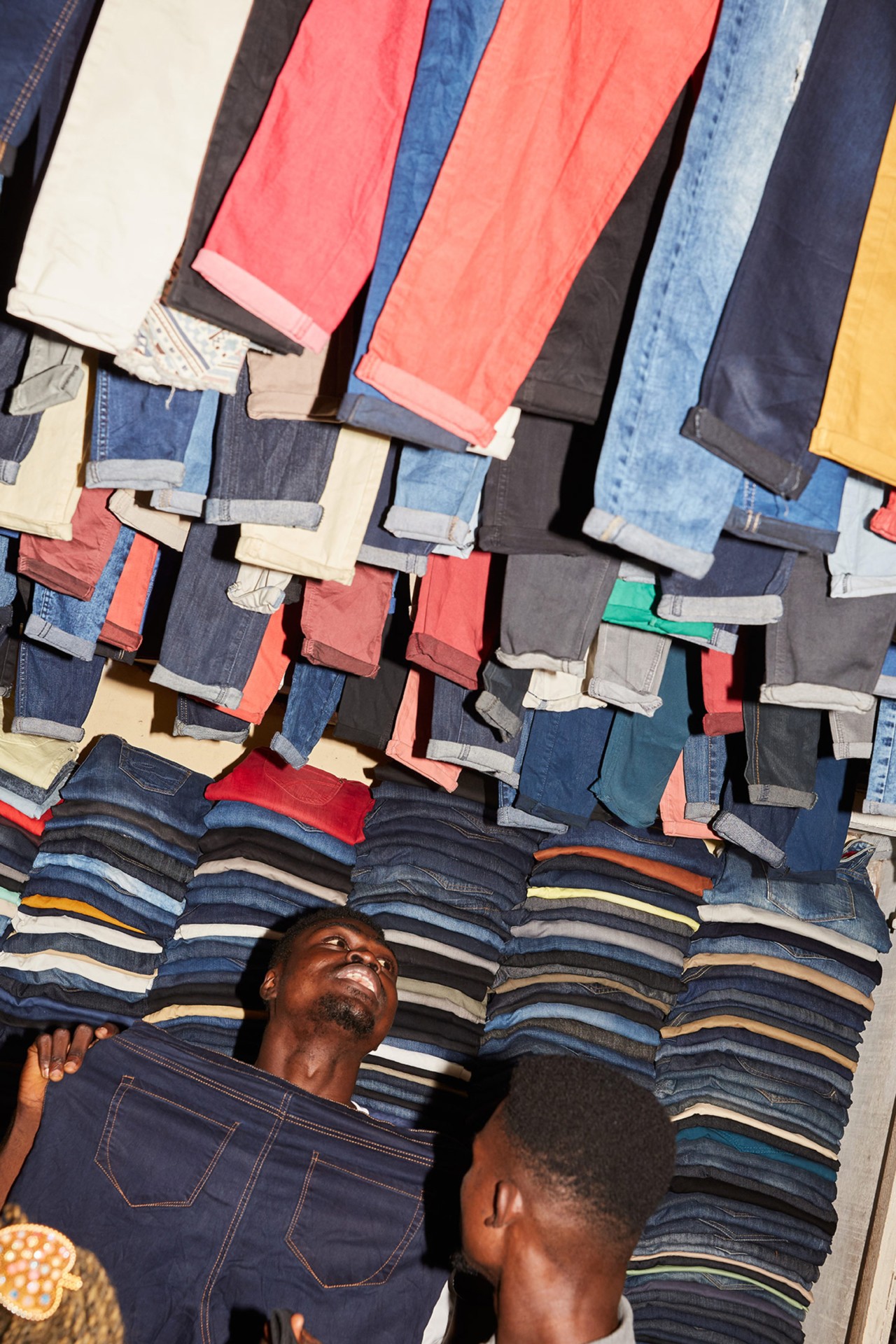 Many jeans are stacked and hung in a retailers stall in Kantamanto market in Accra Ghana by Charlie Engman