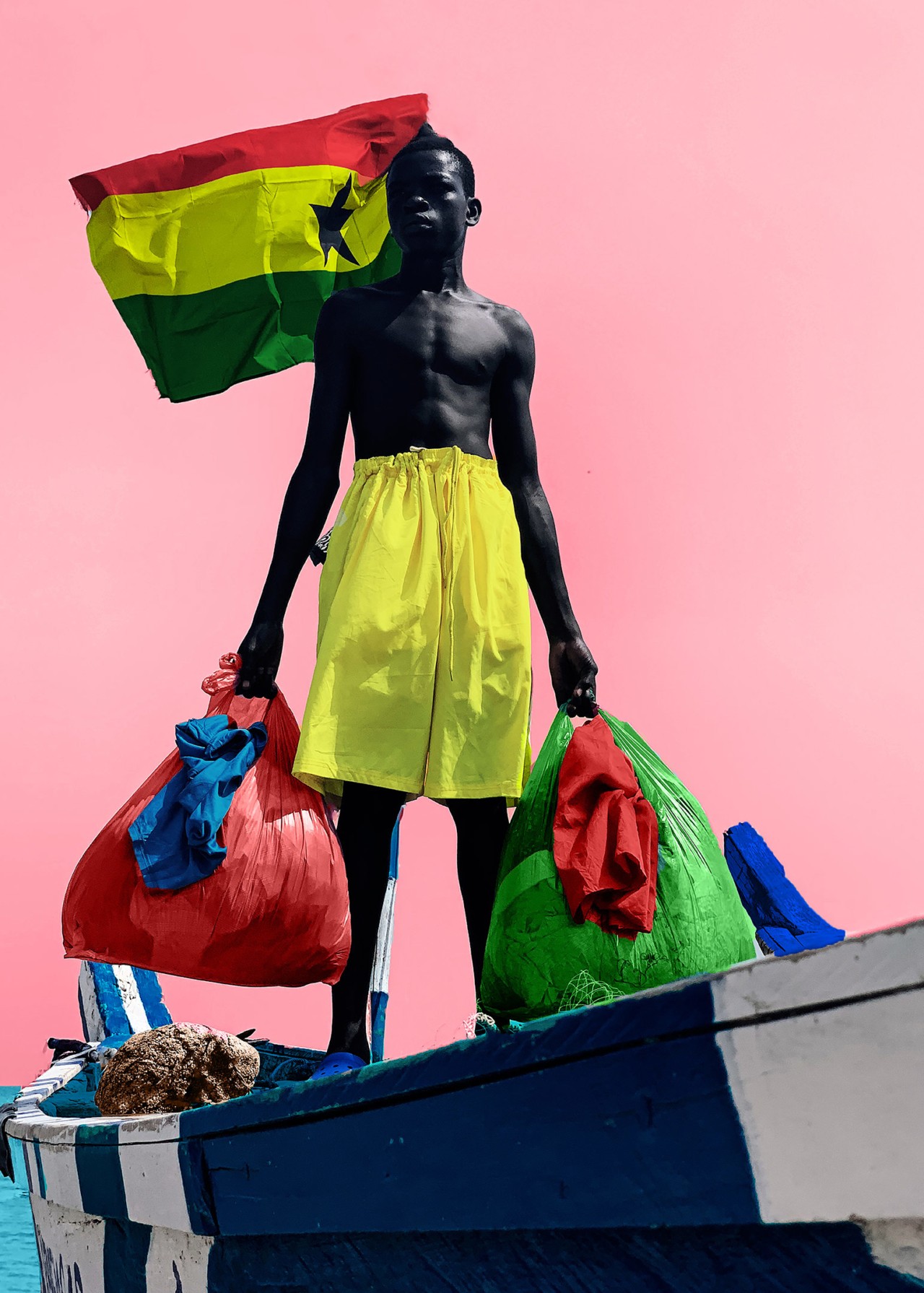 A boy wearing yellow shorts and the Senegalese flag behind him carries a green and a red bag.