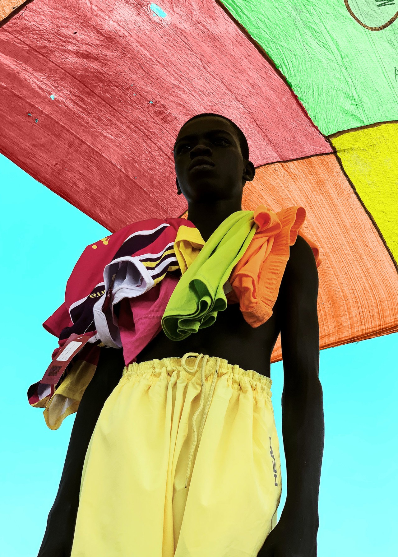 A boy wearing yellow shorts and several garments draped over his shoulders stands below a multi-colored roof.