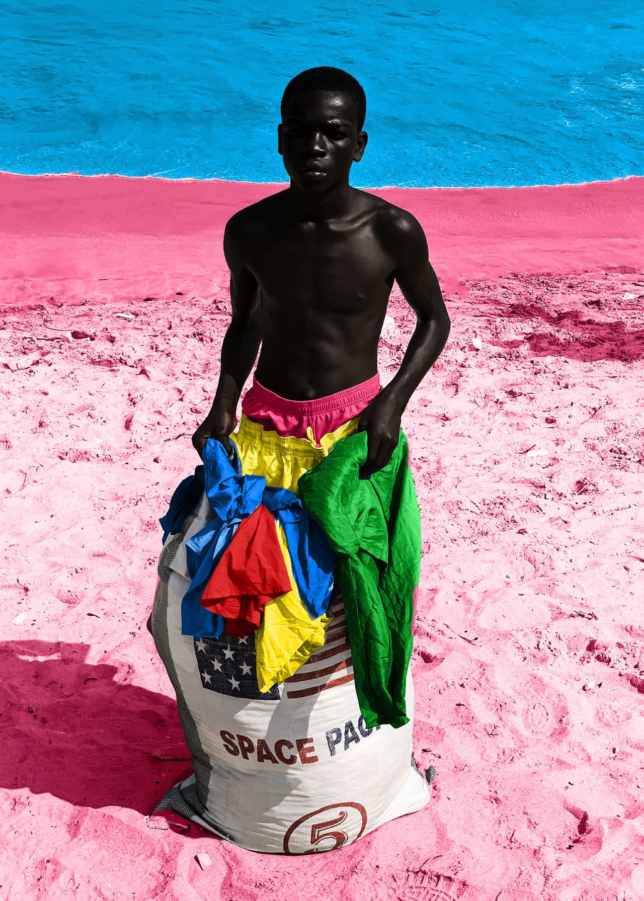 A boy carries a sack full of clothes along a pink sandy beach.
