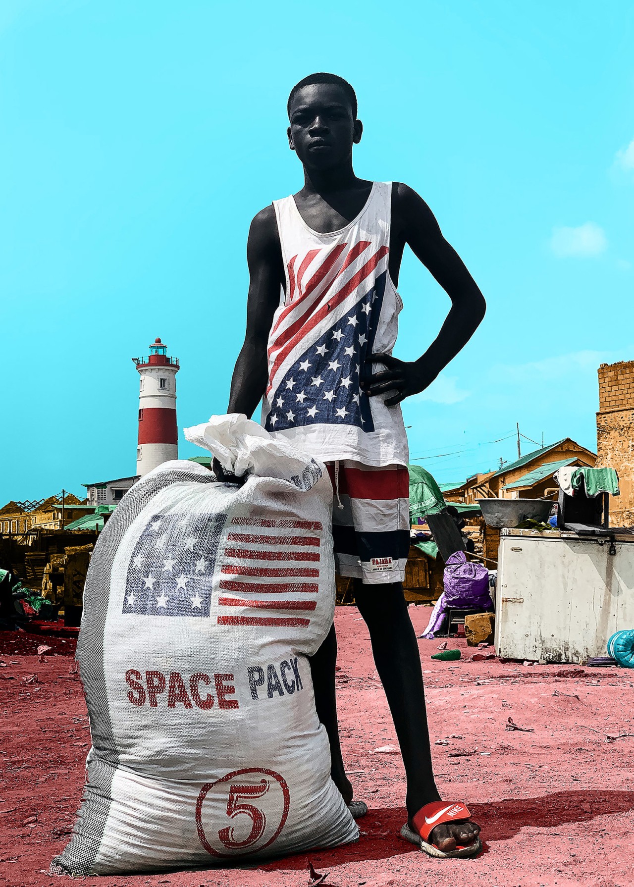 A boy wearing an American flag shirt leans on a large sack of old clothes.