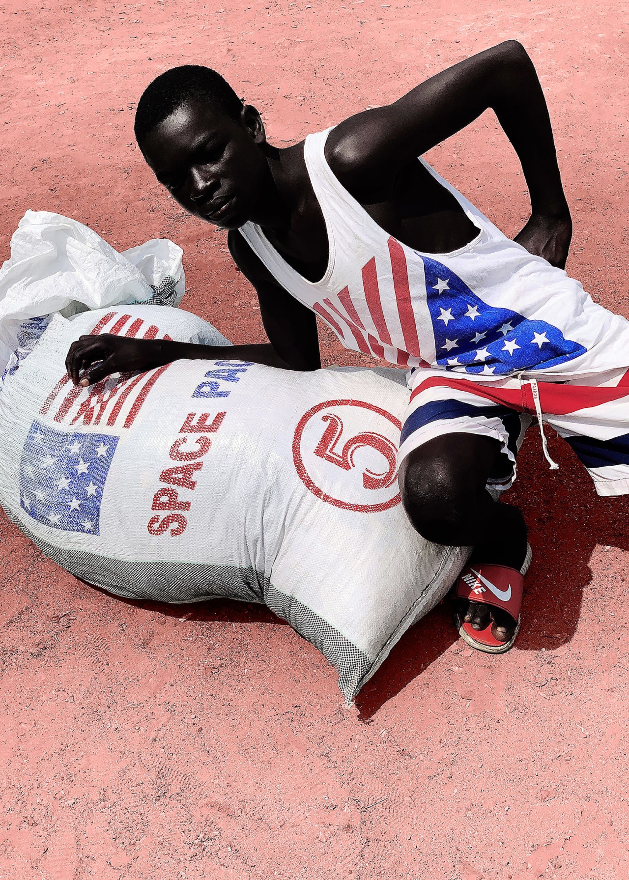 A boy wearing an american flag t-shirt lays down next to a large sack of clothes.