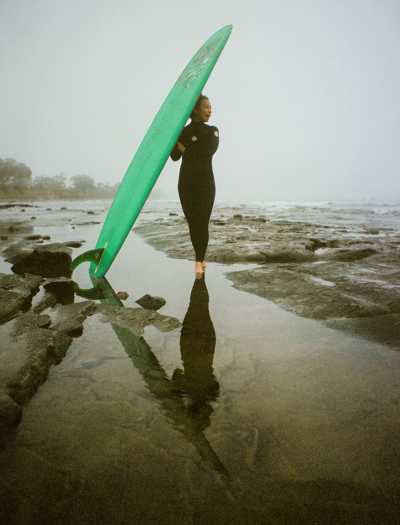 Mixed-race surfer stands in shallow water surrounded by rocky terrain at a California beach. She is holding up a large green surfboard as she looks away toward ocean.