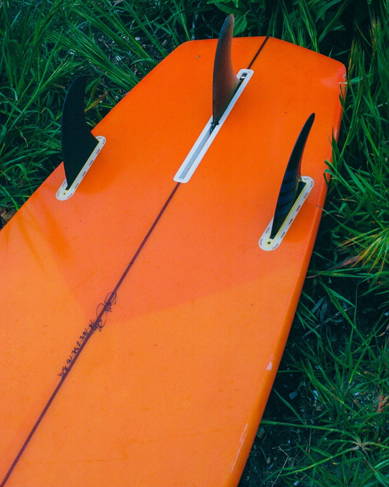 The tail end of a bright orange surfboard lies still on a bed of grass.