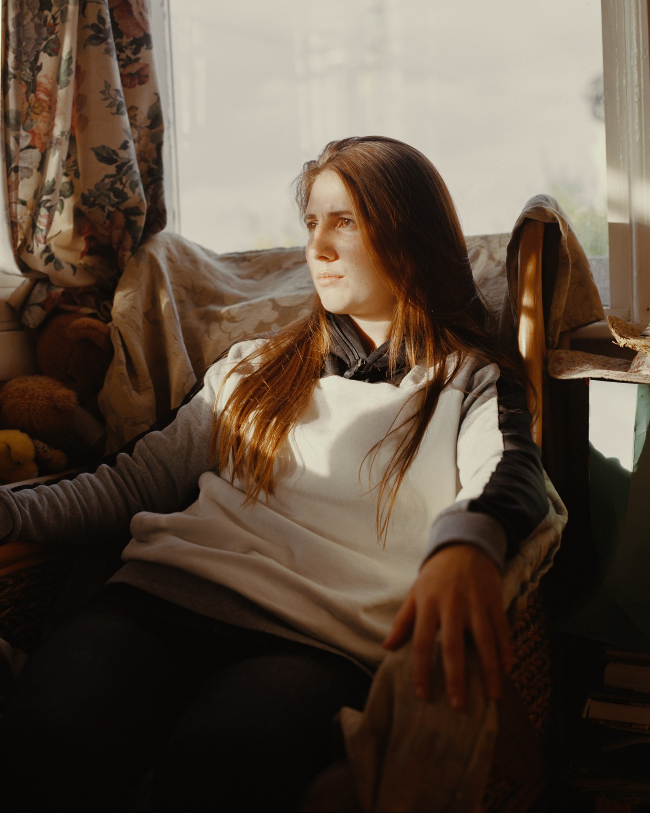 A woman sits in a chair in her dimly lit British east coast home. Behind her is a large, opaque window with flower decorated curtains bunched to the side.