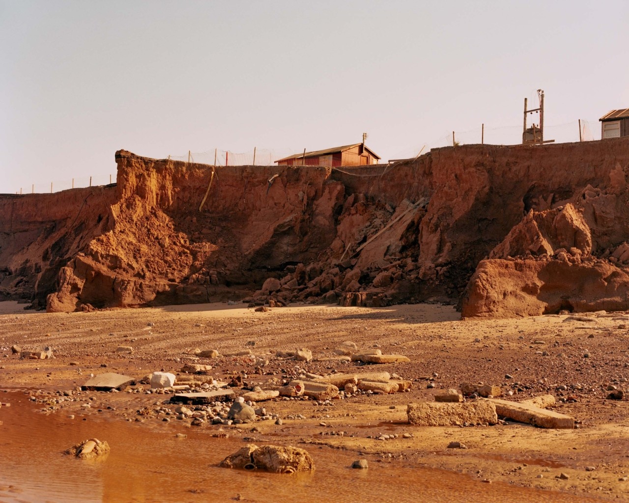 A house sits along the edge of a jagged, eroded cliff, surrounded by a metal gate and power line. Below cliff, water washes up on the rocky, yet flat and sandy shore.