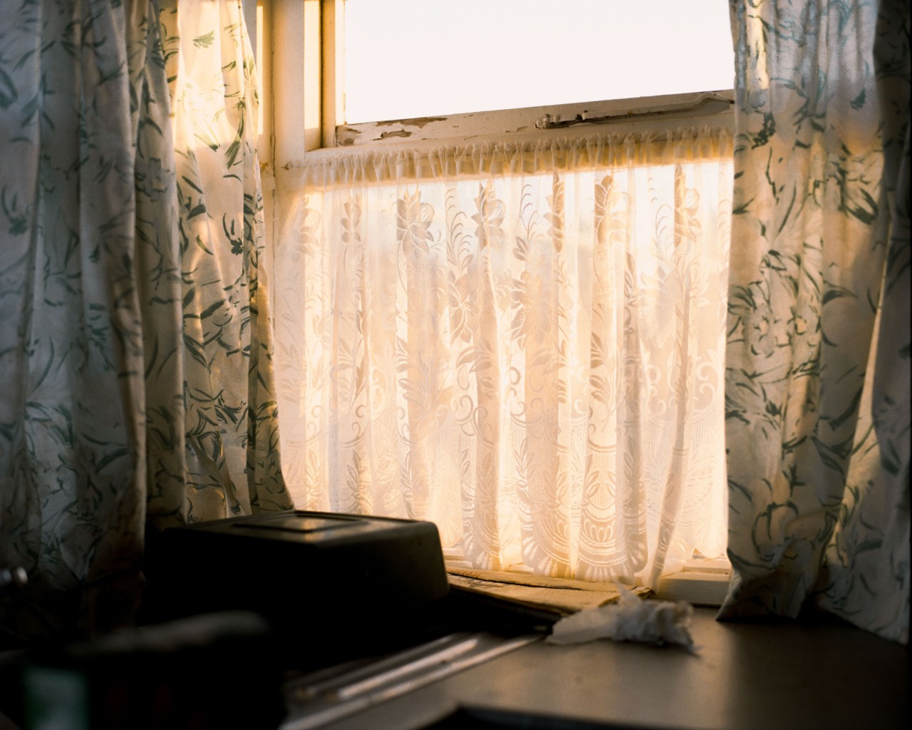 Sheer white, flower decorated curtains cover a glass window while the sun barely shines through. Larger green and yellow flower decorated curtains frame the window on both sides.