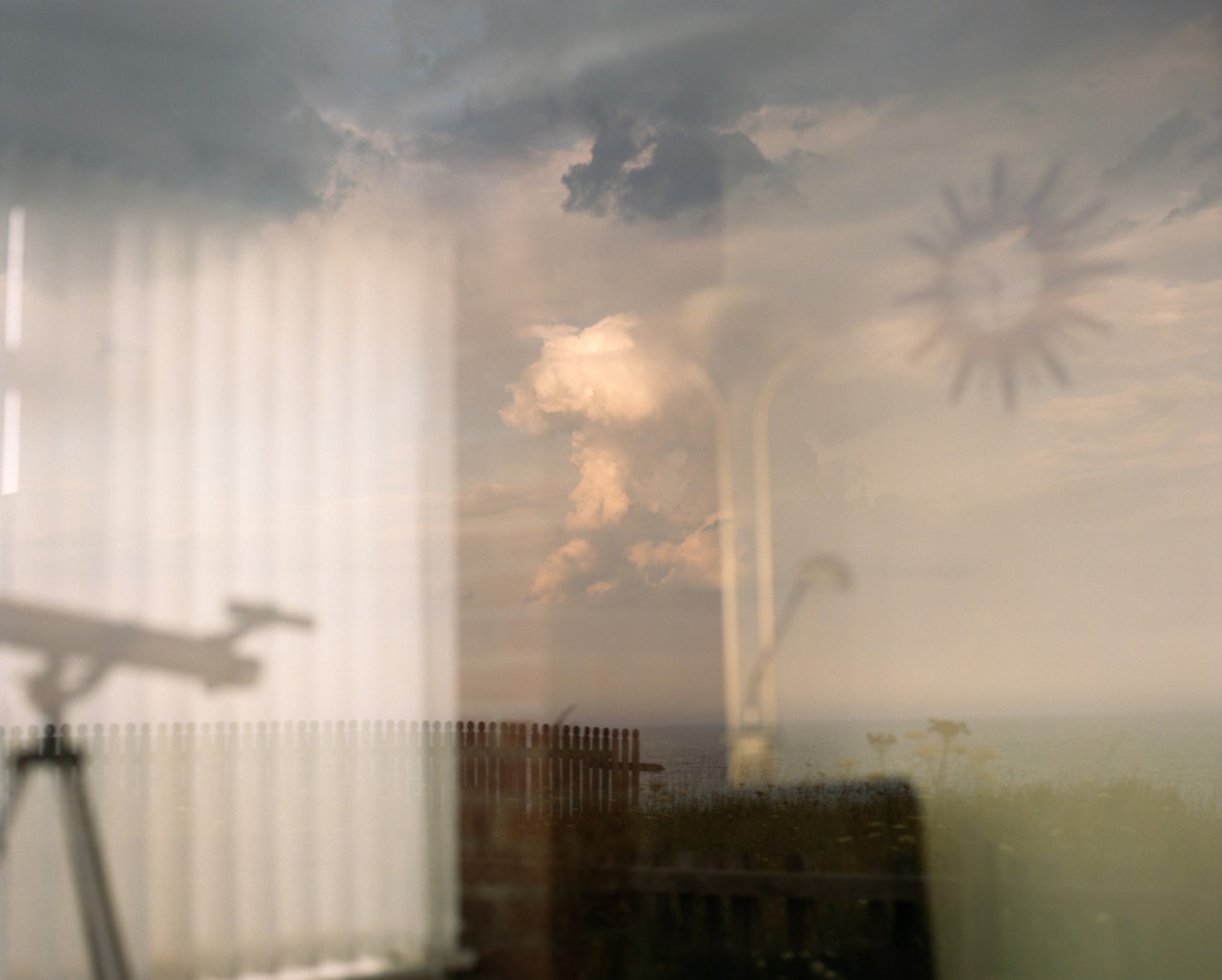 A cloudy sky, ocean, and wooden gate is hazily shown through a glass window. In the reflection is the inside of a home with a clock on the wall, blinds, and a telescope.
