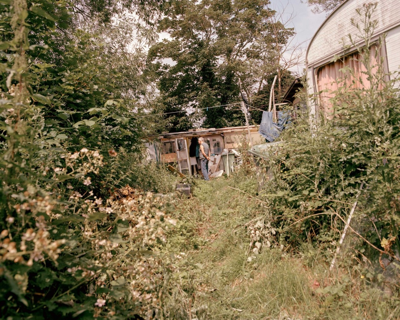 A man is framed by foliage, standing in the distance in front a mobile home surrounded by forest.