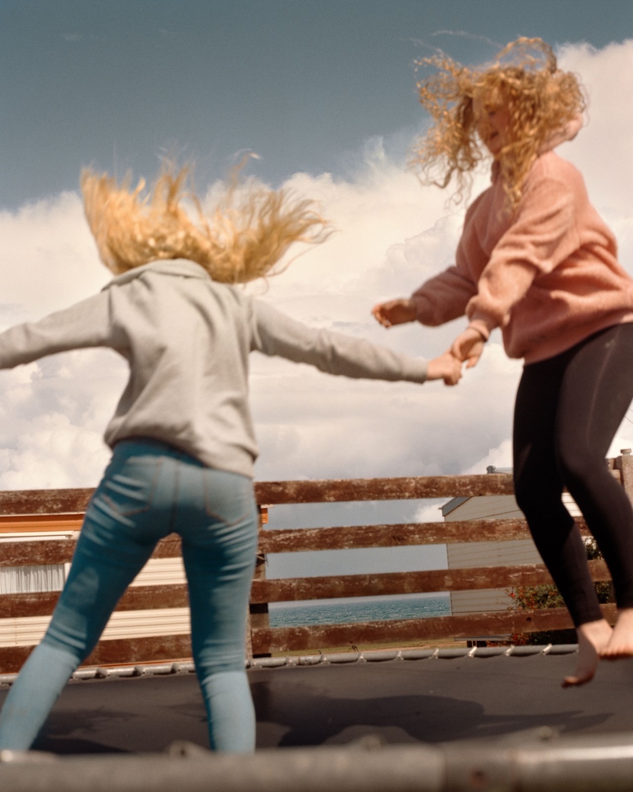 Two young, blonde adults hold hands while jumping on a trampoline. A brown wooden gate separates them from the British coastline.