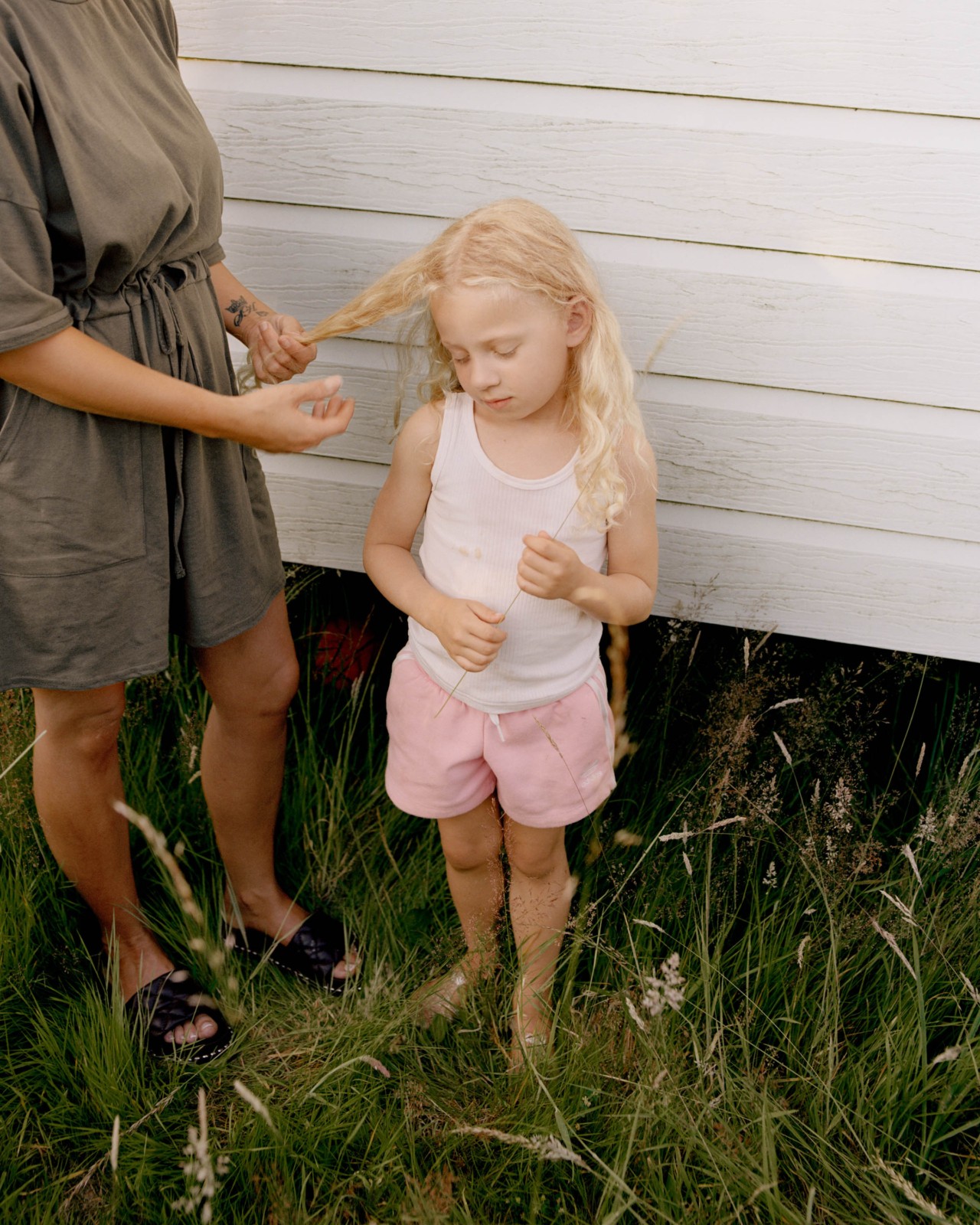 A woman with her face not shown, stands next to a small, blonde girl on a patch of grass in front of wooden white blocks. The young girl looks down while the woman plays with the girl's hair.