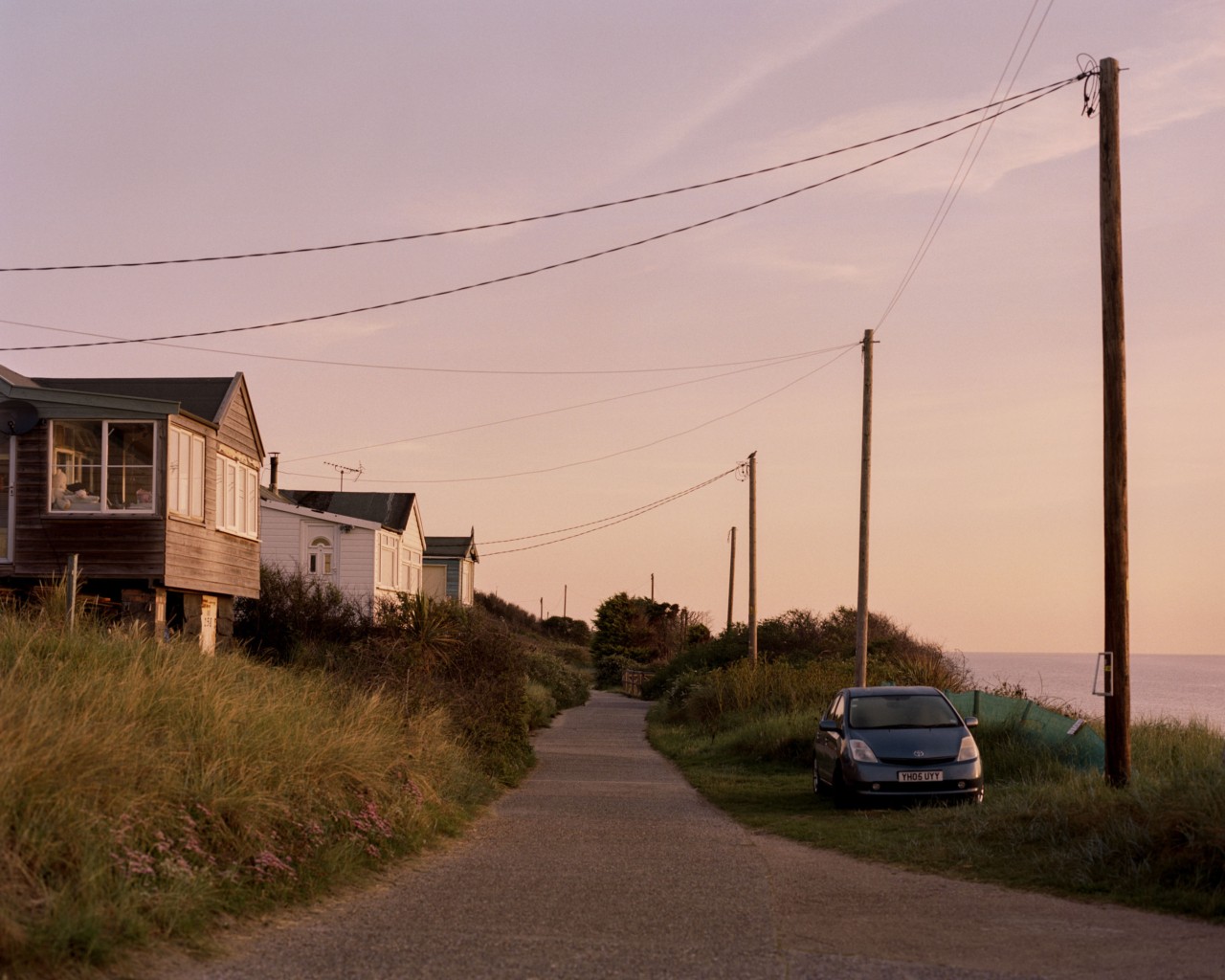 A road is framed by houses and foliage on one side, the British coast on the other, and power lines and a sunset sky above, in this British east coast community.