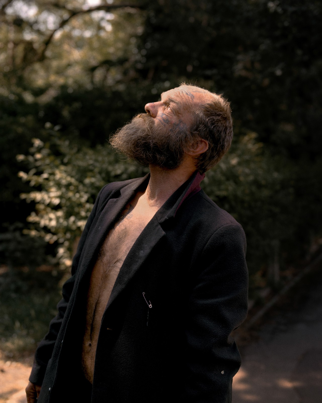An older man stands in front of a foresty background looking up toward the sky.