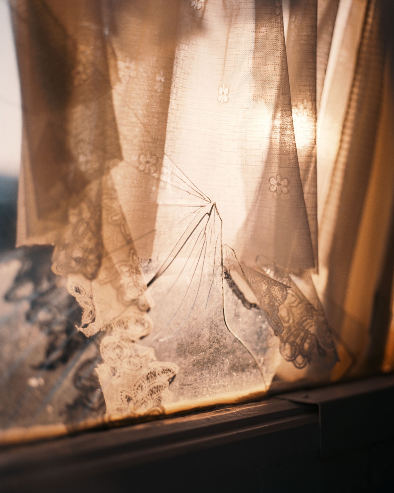 The sun peeks through a cracked glass window facing the sea. Window is slightly concealed by sheer, decorative white curtains.