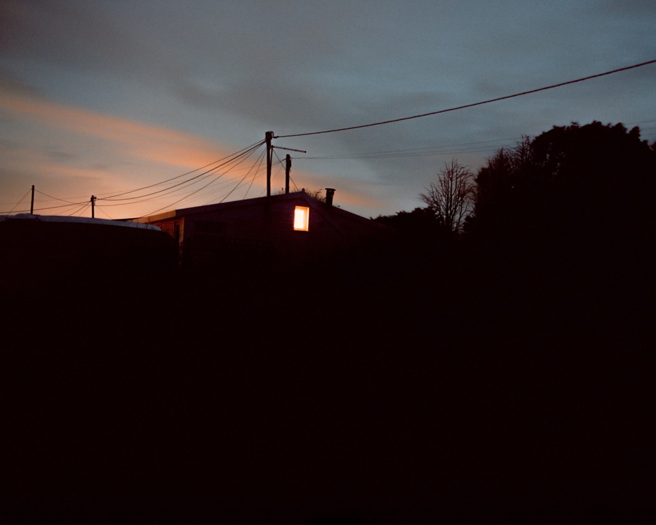 The silhouette of a house sits in the distance with one brightly lit window at the top. Dark shadows are in the forefront, while a power line is shown above the house and below a blue-ish pink sunset sky.