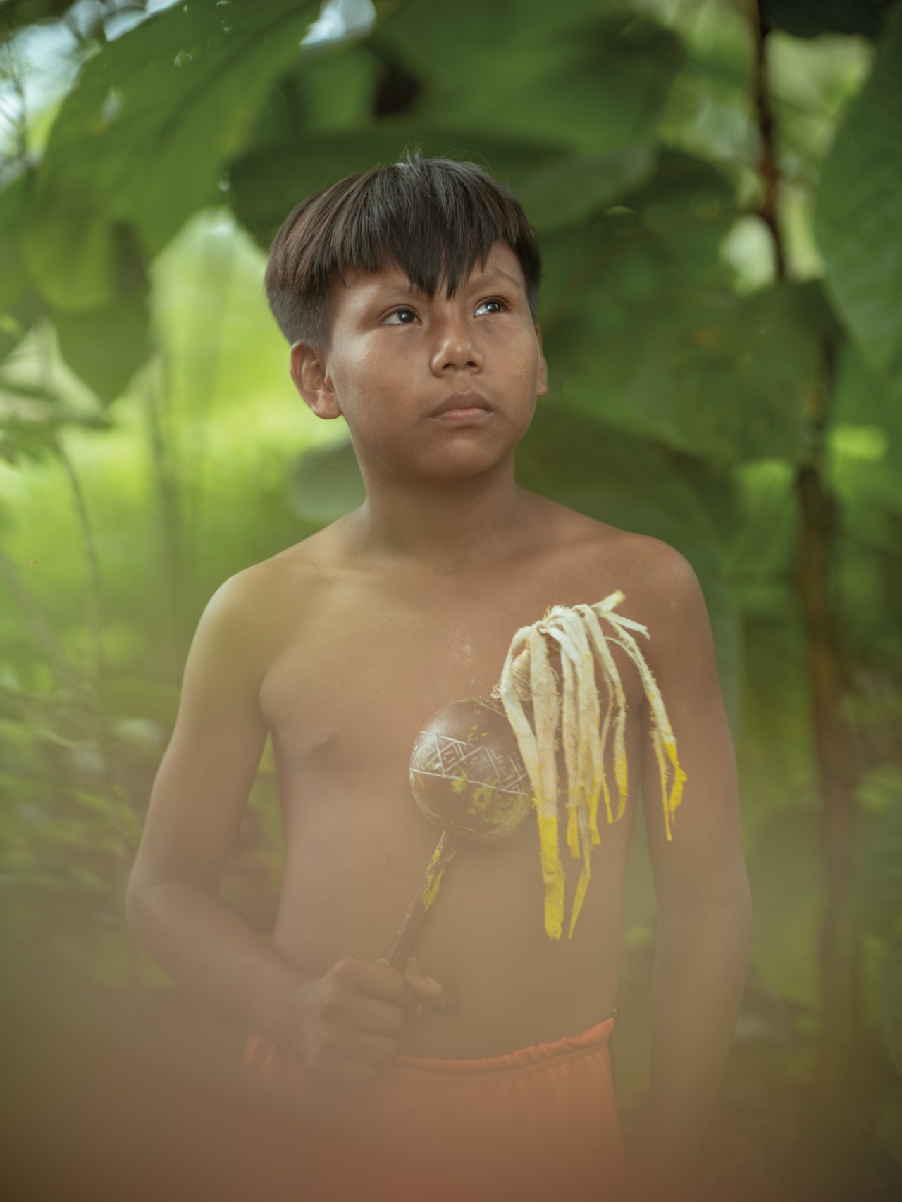 A young boy stands in the Amazon rainforest, looking toward the sky while holding a home-made instrument in his right hand.