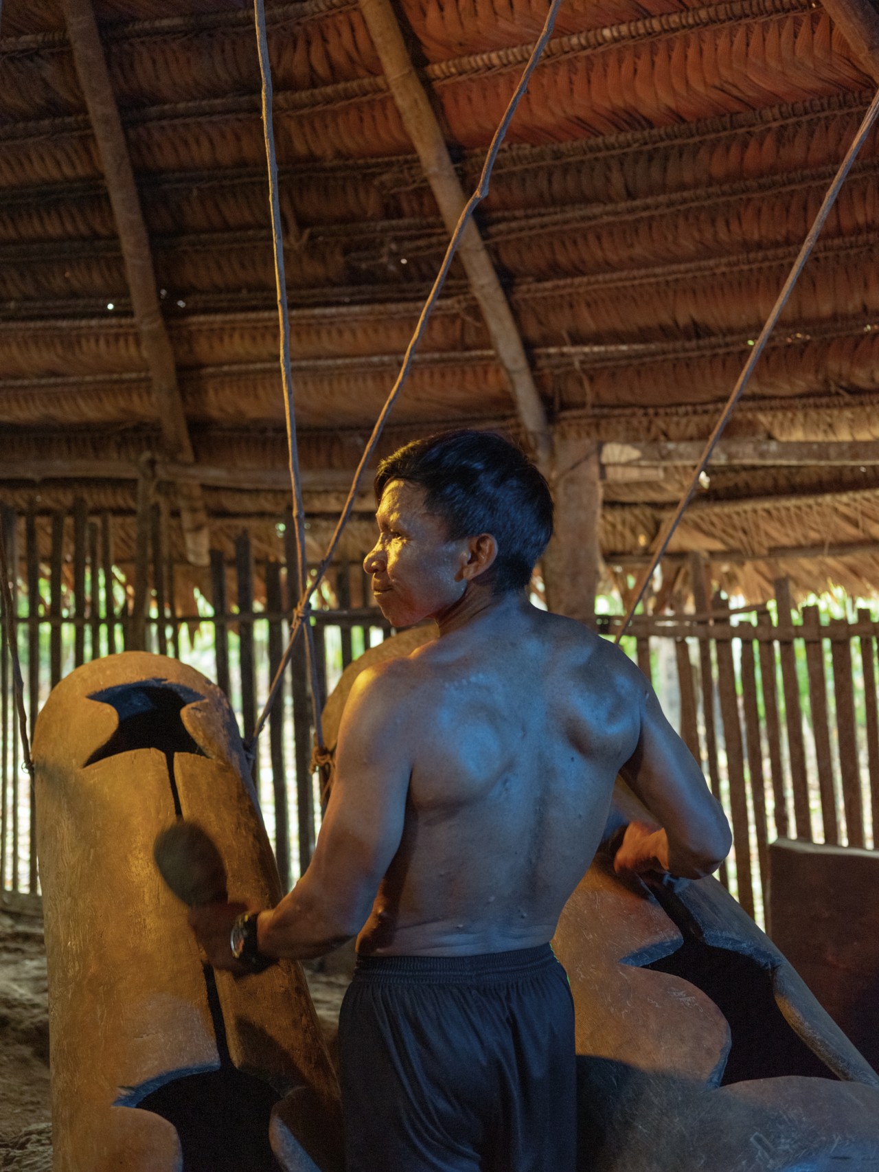 A man faces away from the camera while beating on two large drums in an Amazon rainforest shelter.