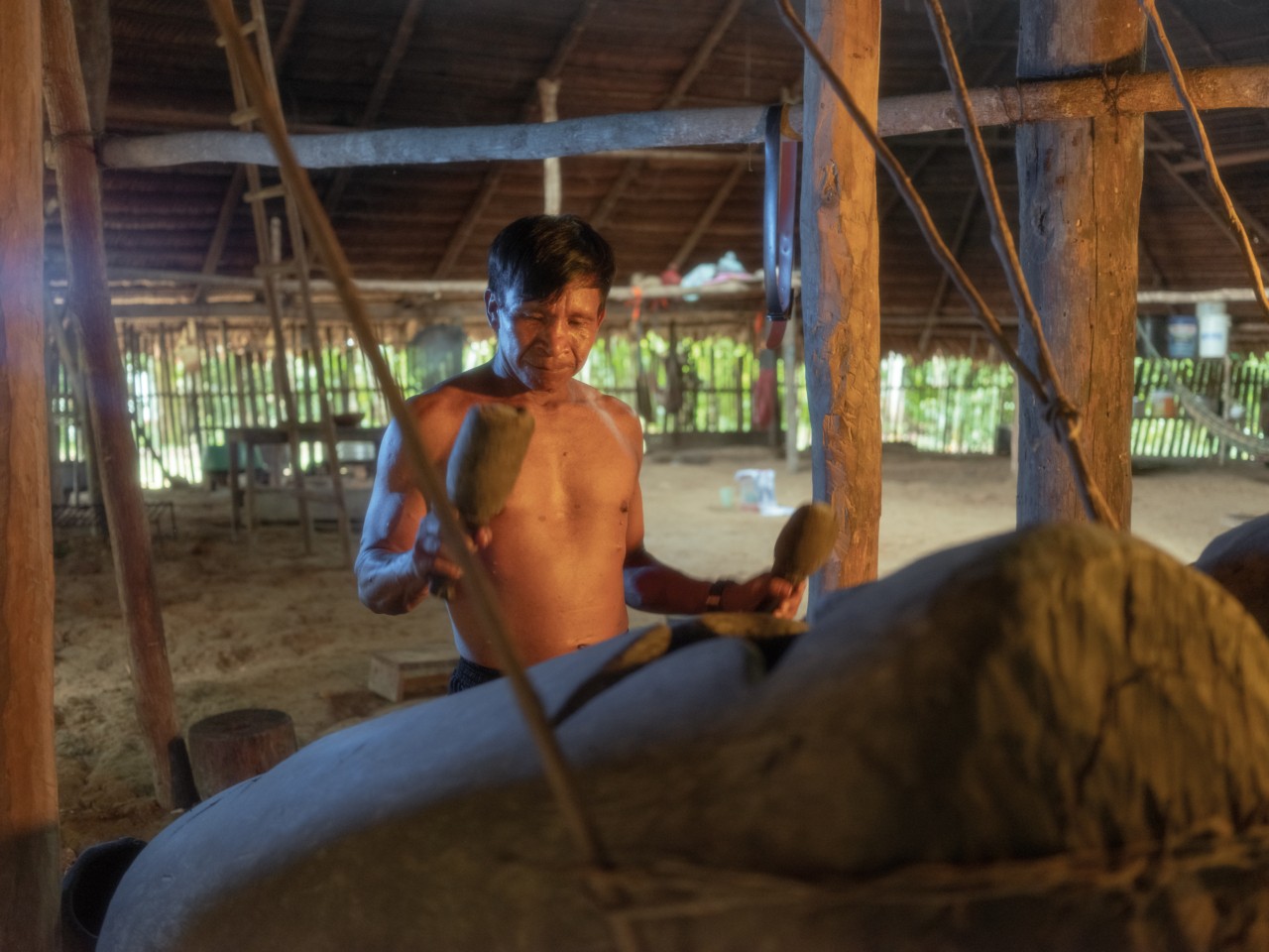 A man stands inside an Amazon rainforest shelter beating on a large drum.