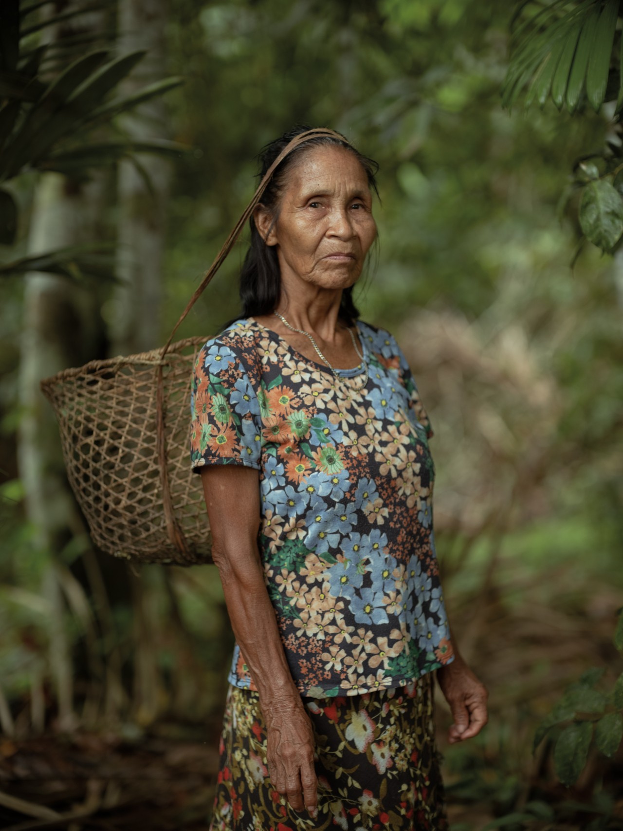 An older woman looks toward the camera while standing in the Amazon rainforest. She has a basket hanging from her forehead by a thick strap.