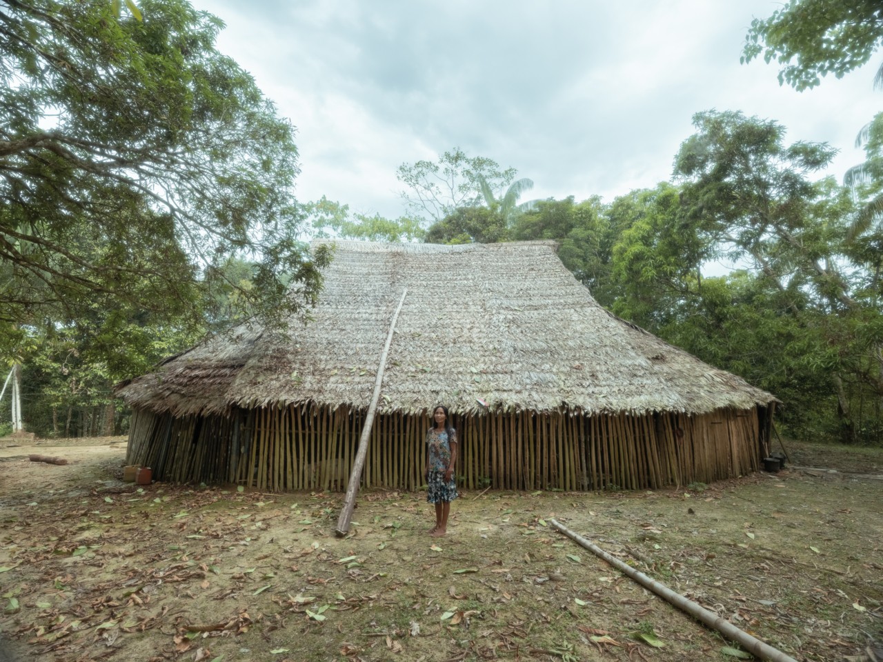 A woman stands in front of large hut that sits in the center of a clearing in the Amazon rainforest.