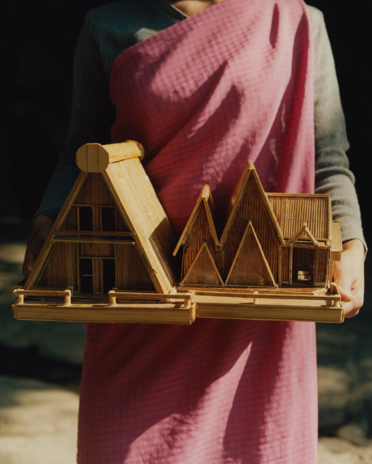 A woman wearing traditional Indian garb is holding a small replica of a wooden house. Her face isn't shown.