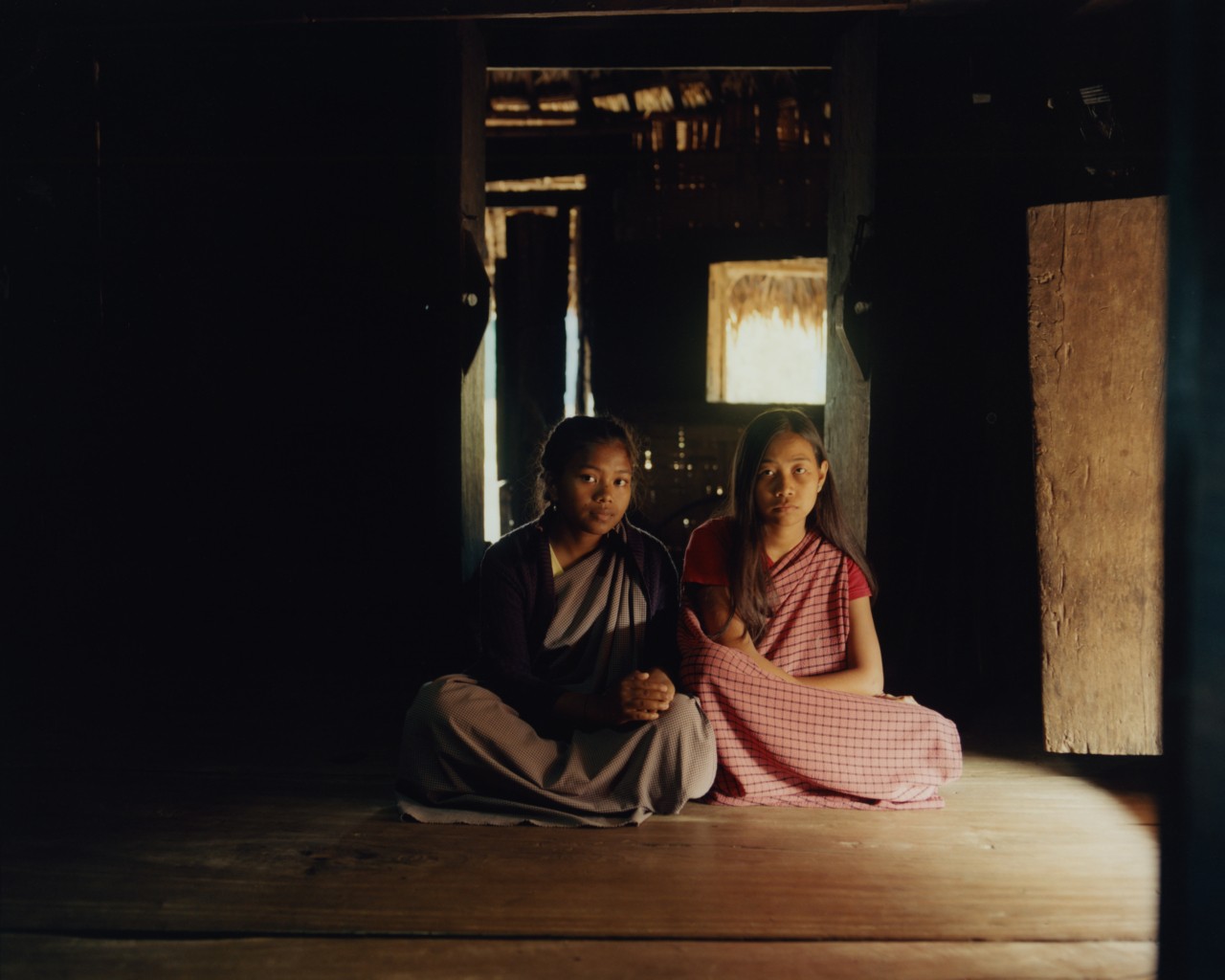 Two young girls sit with their legs crossed on the floor of a house.