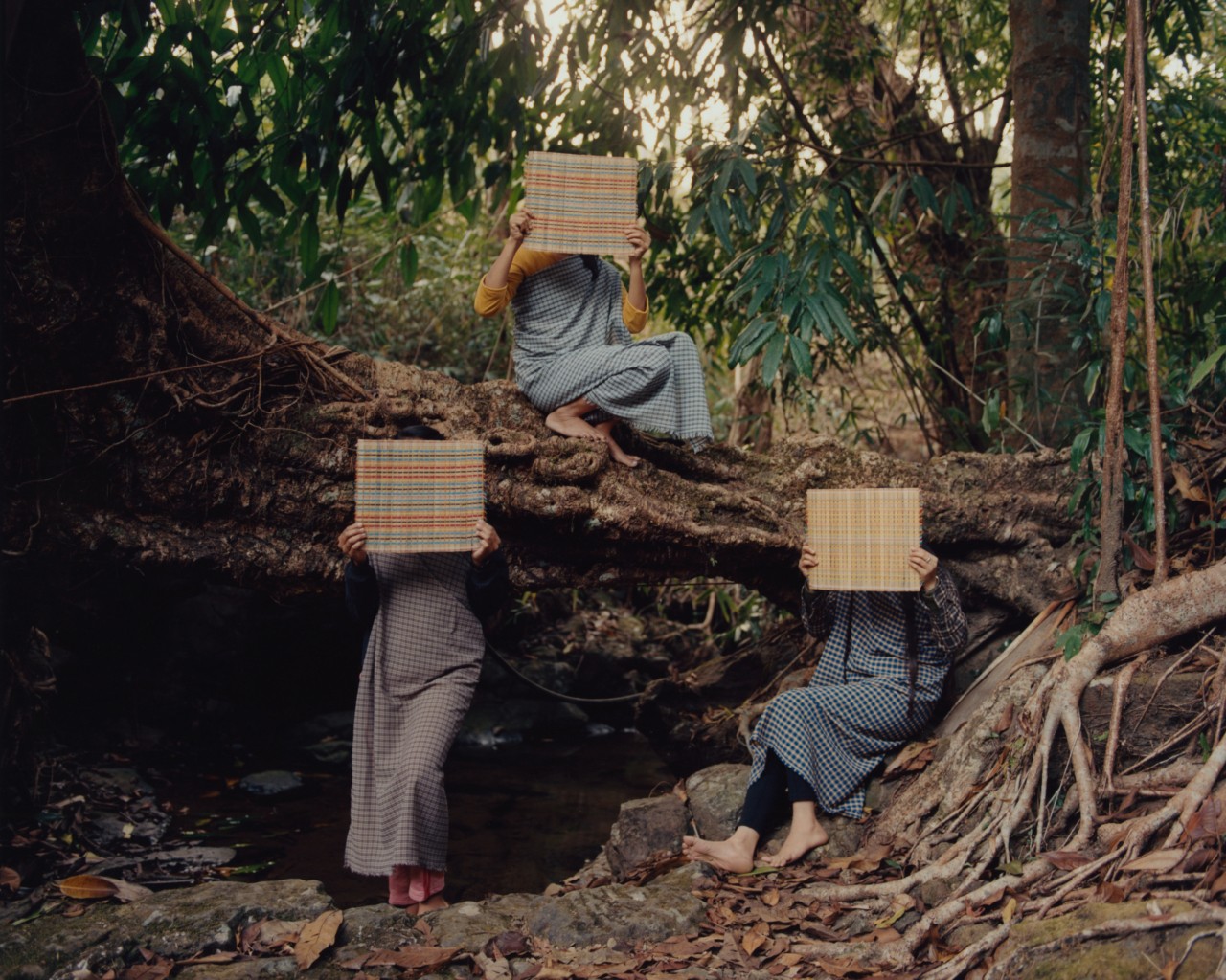 Three people cover their faces with a square woven basket while sitting and leaning on a large tree in the ancient forest.
