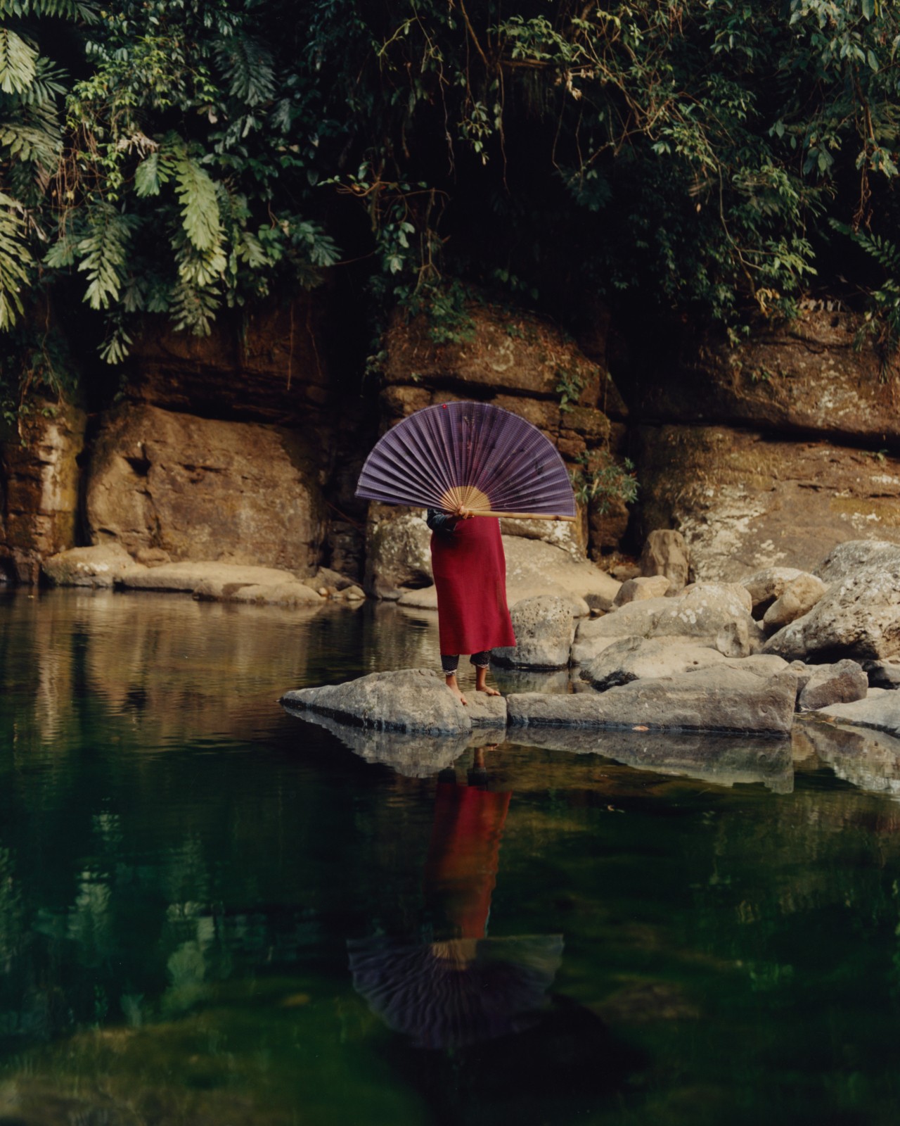 A person stands on a rock in the middle of a body of water, while covering their face with a large purple fan.