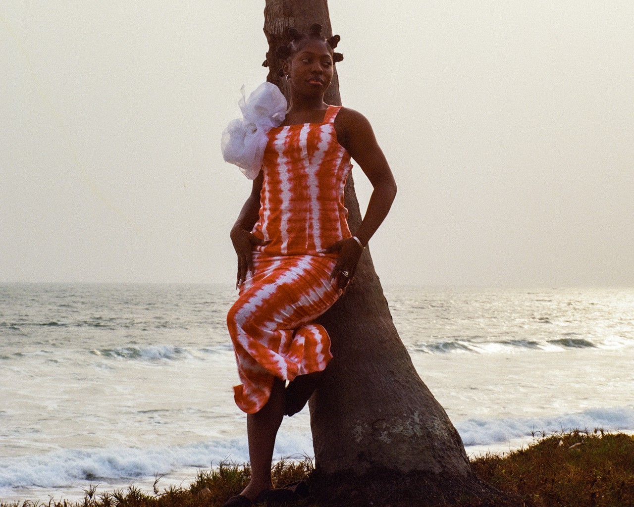 A young woman leans against a tree along the coastline.