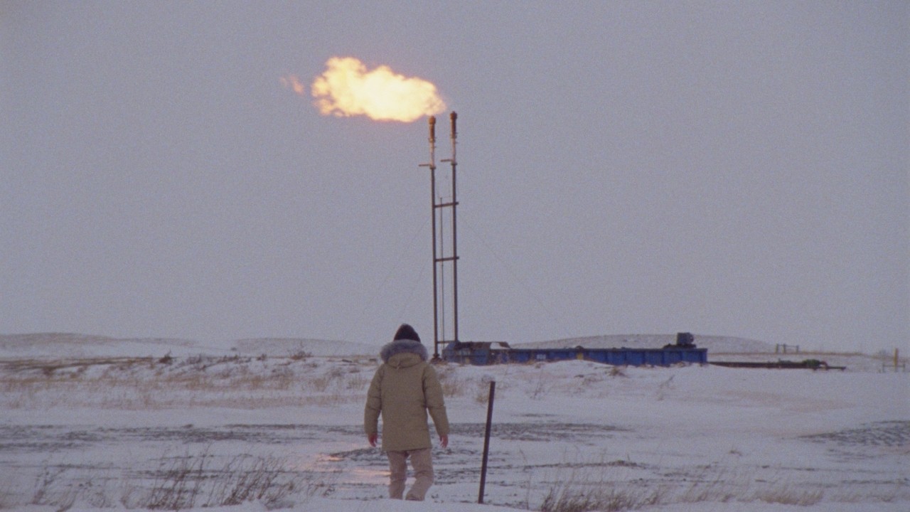 A scene from Daniel Goldhaber's How To Blow Up A Pipeline movie. A man facing away from camera walks on a snowy field toward a pipeline explosion in the distance.