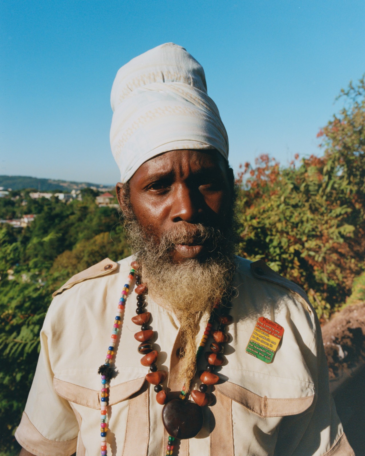 A Jamaican man with his hair wrapped into a scarf and a pin of the Jamaican flag on his shirt looks straight into the camera. The Blue Mountains are in the background.