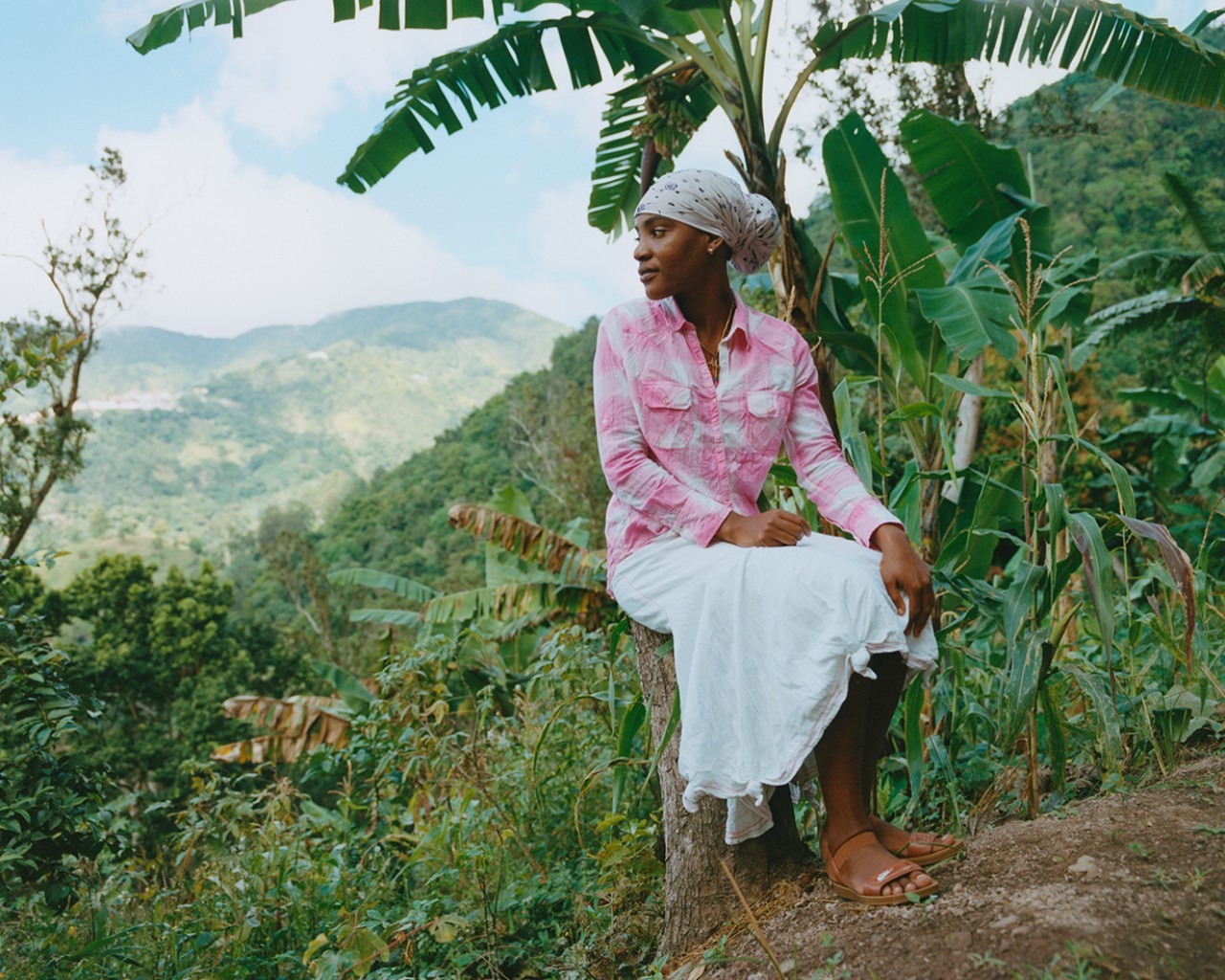 A Jamaican woman with her hair tied into a scarf sits on a tree stump in the middle of the Blue Mountains.