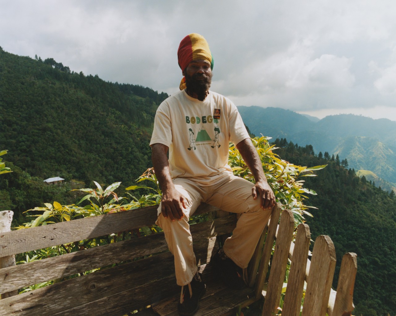 A Jamaican man with his hair wrapped in a red, green, and yellow scarf sits on top of a bench with the large Blue mountains and a cloudy sky in the background.