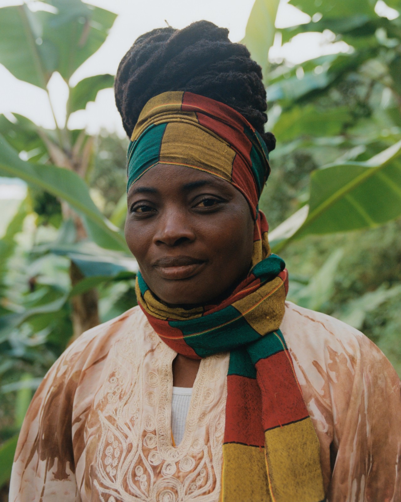 A Jamaican woman wearing a yellow, red, and green scarf looks toward the camera as she stands in the jungles of Jamaica.