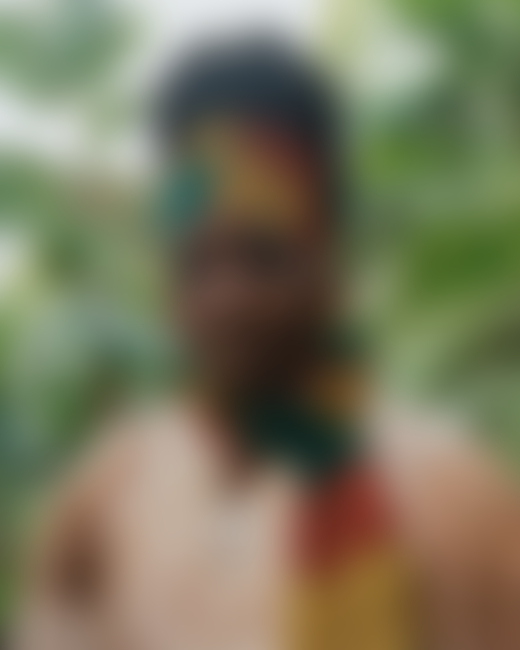 A Jamaican woman wearing a yellow, red, and green scarf looks toward the camera as she stands in the jungles of Jamaica.