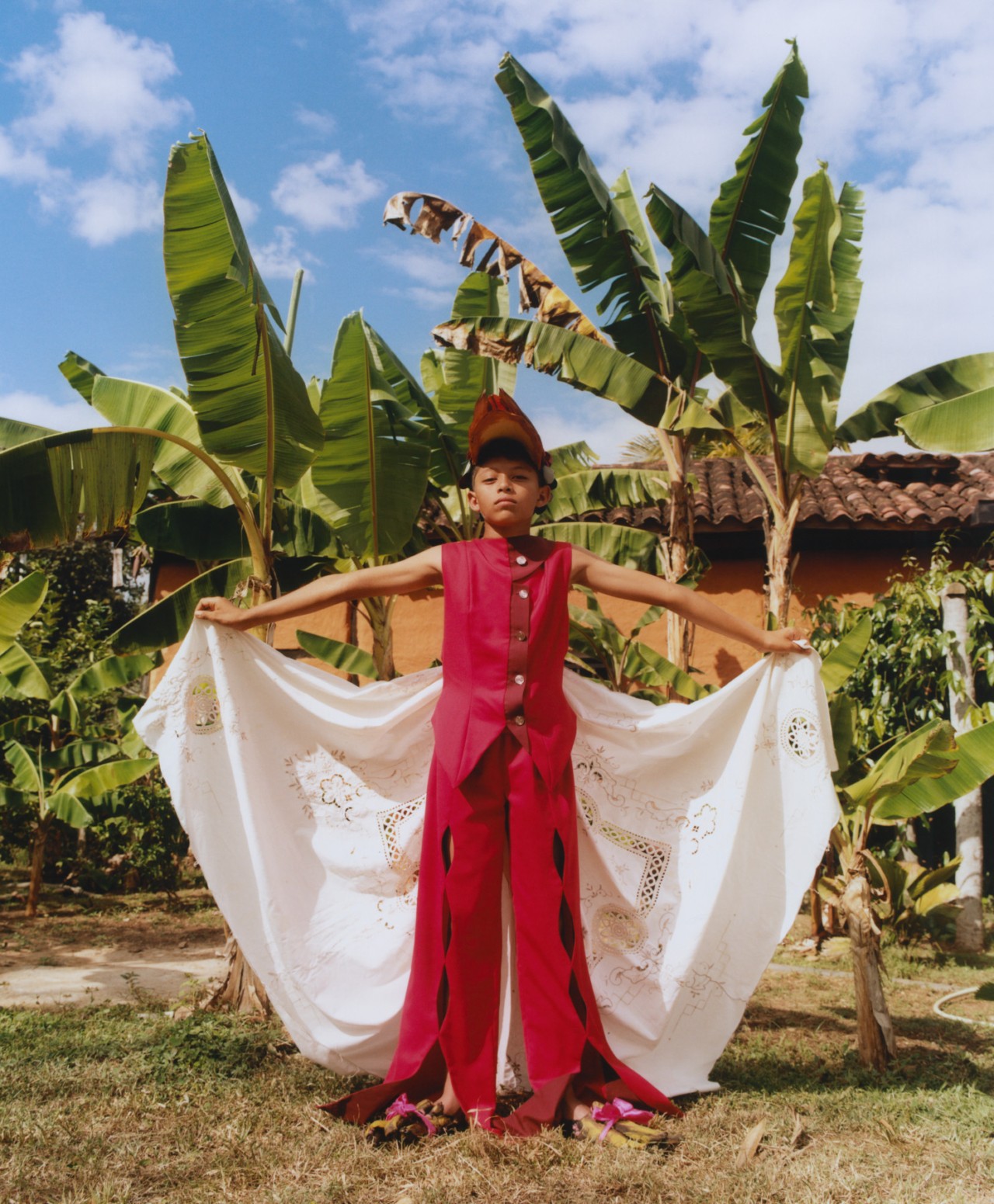 A young Venezuelan boy stands in front brightly painted Venezuelan home with large plants in front of it. His arms are stretched out to the sides, holding onto his garment.