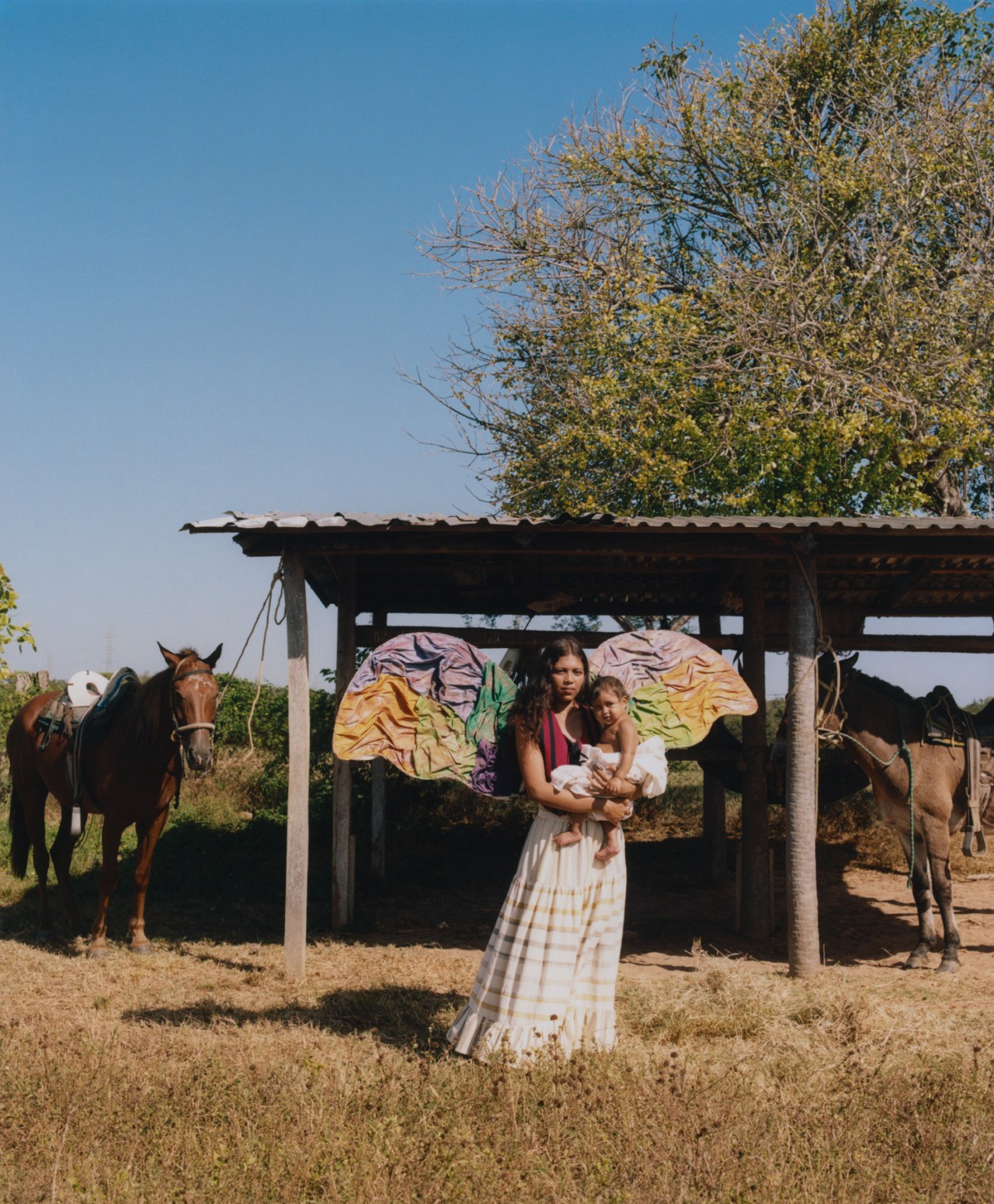 A young Venezuelan woman holding a baby stands in front of a horse stable. She has colorful wings attached to her back.