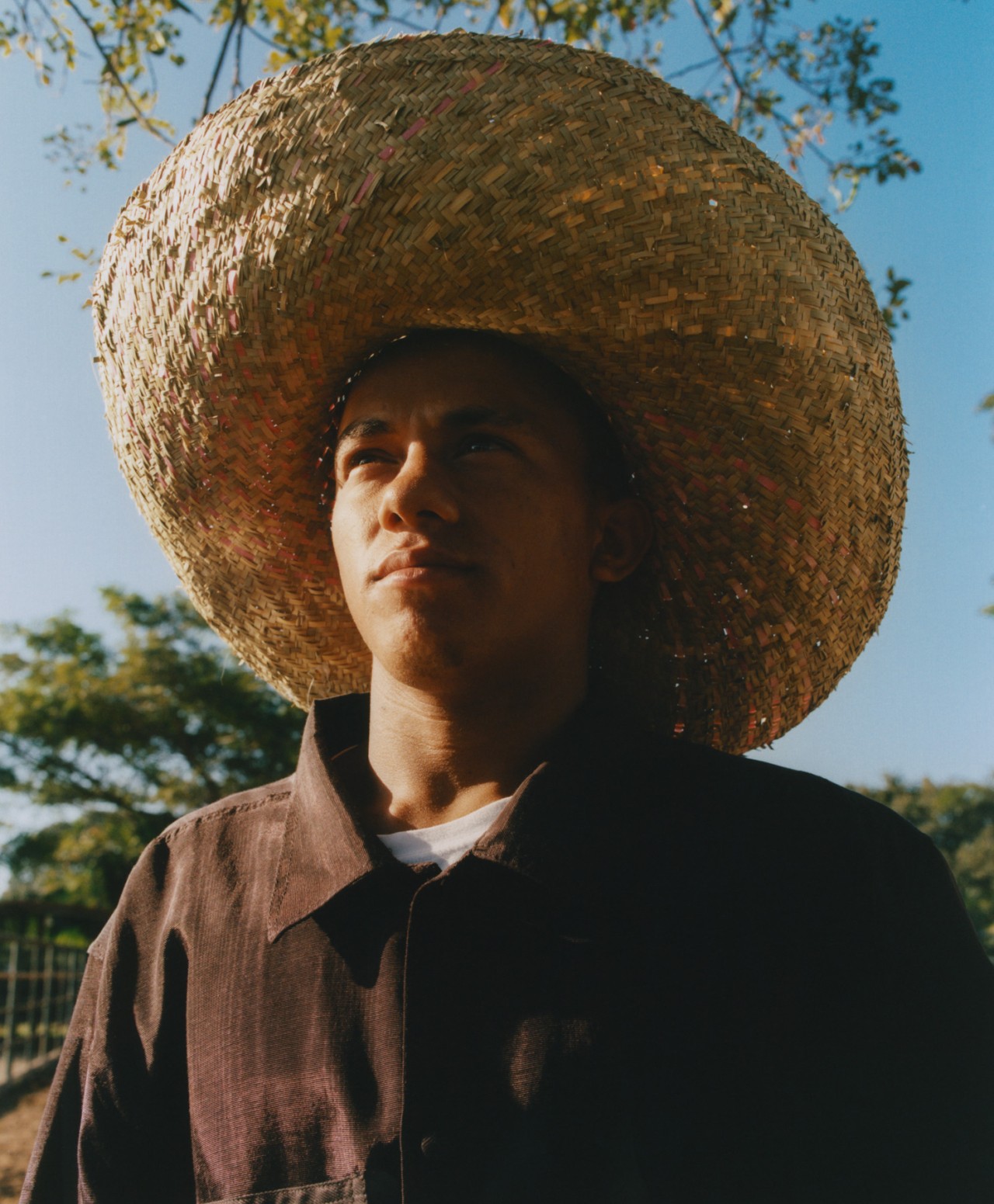 A young Venezuelan man wearing a large straw hat and a brown collared shirt looks up toward the sky.