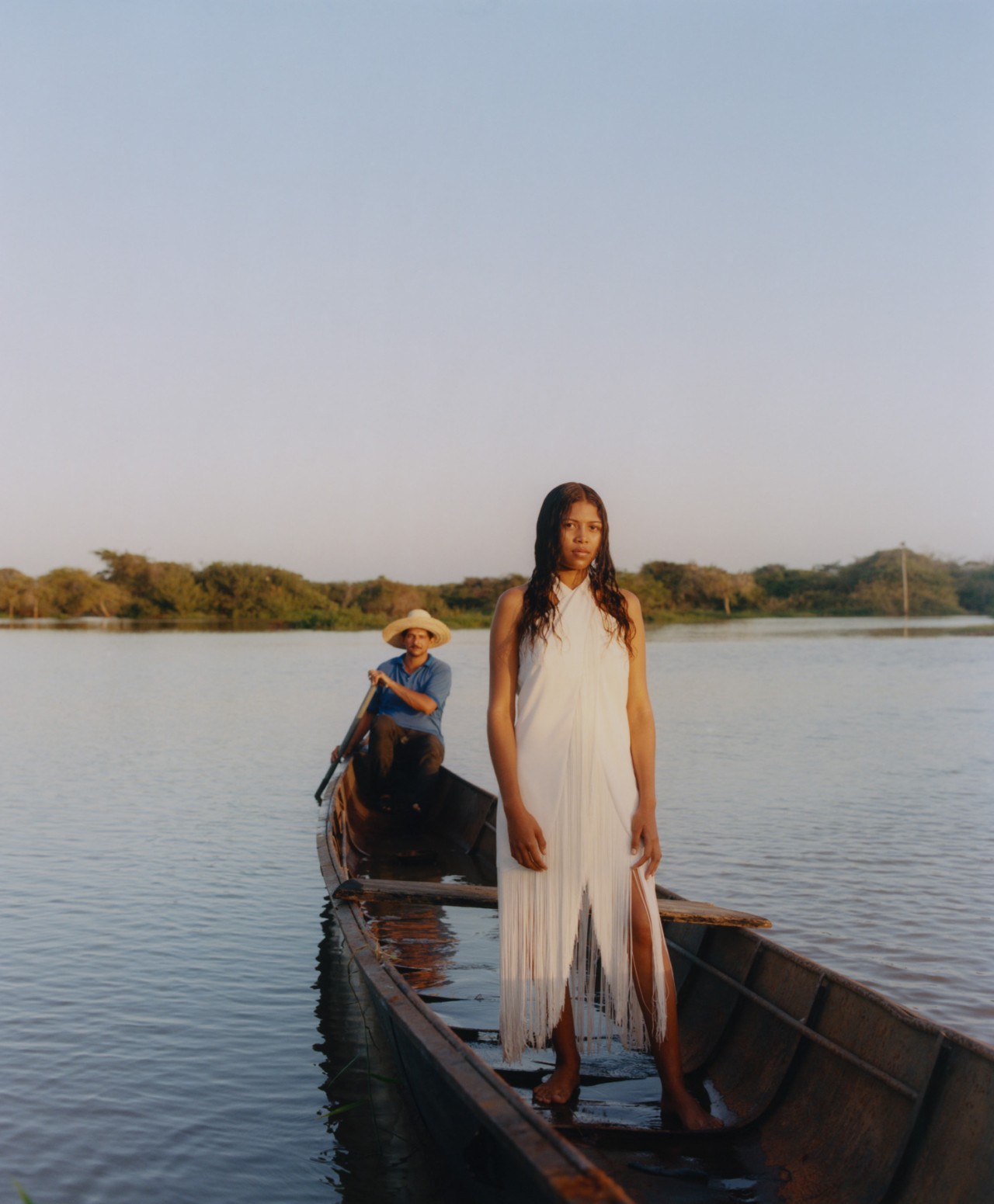 A young Venezuelan woman in a white dress stands up inside a boat with a man sitting behind her rowing them through the water.