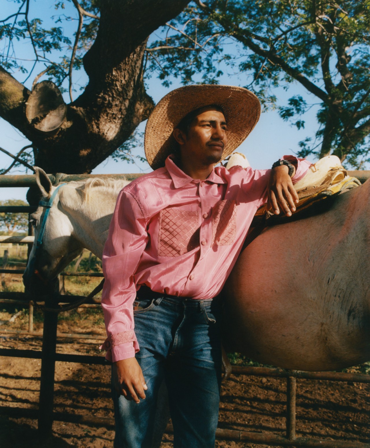 A young Venezuelan man dressed in a straw hat, a collared shirt, and high waisted jeans leans against his horse in a stable in Venzuela.