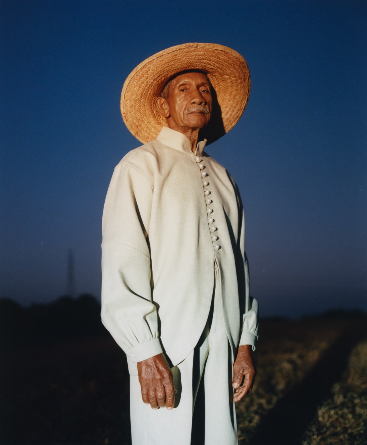An older Venezuelan man stands amid the Venezuelan plains staring straight ahead.