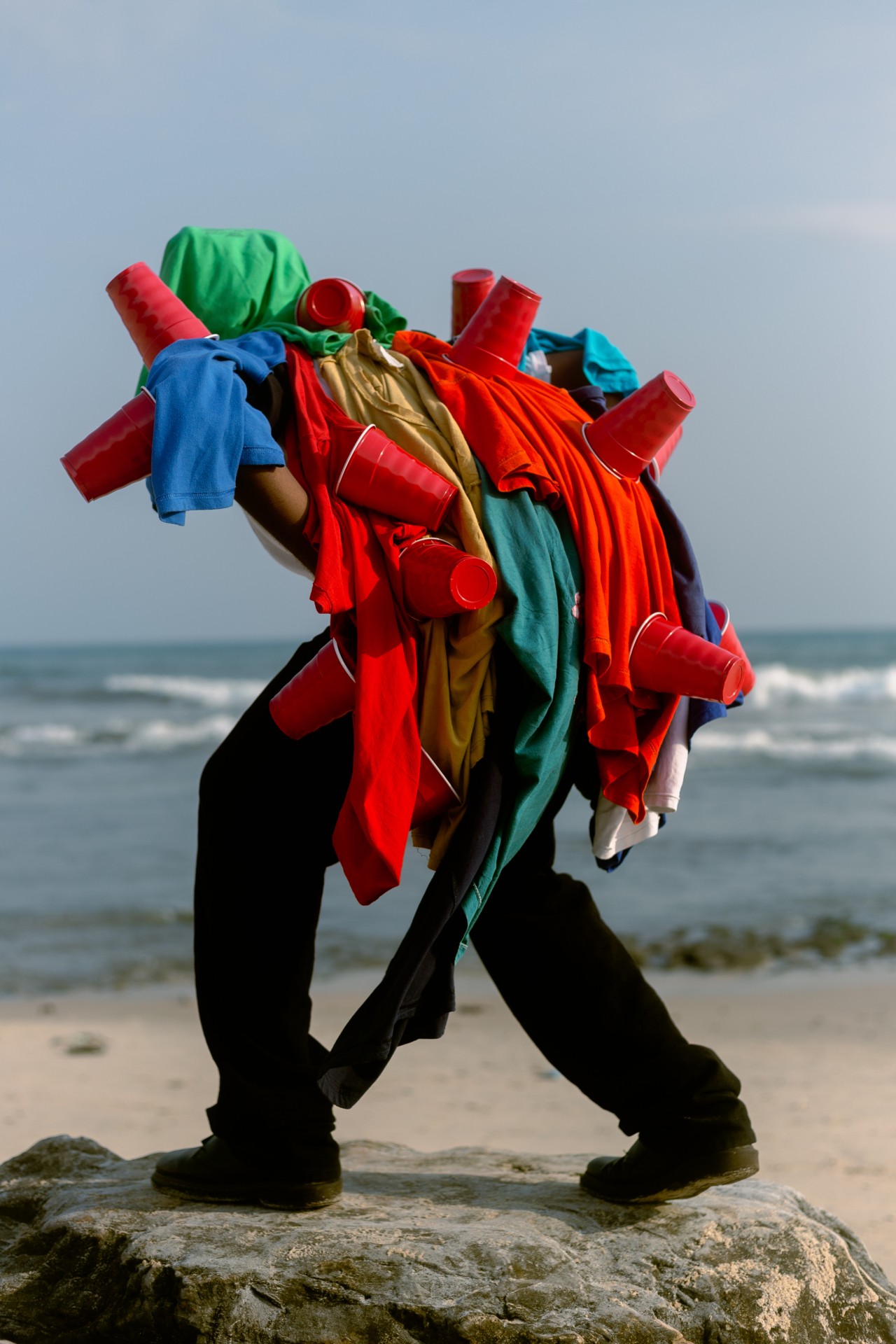 Someone stands with their back crouched over, on a rock on a beach. They are covered in different colored clothing and red plastic cups