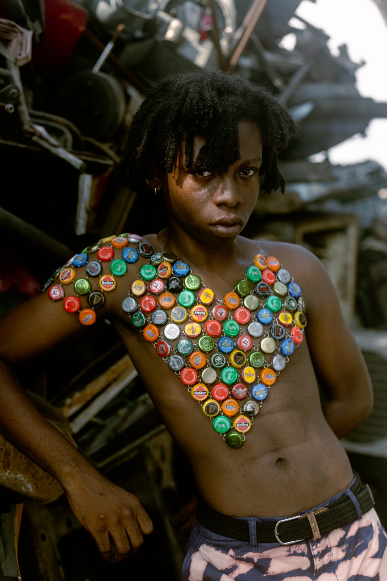 A young man poses in front of a pile of junk. He is shirtless with an array of aluminum bottle tops arranged in a heart on his chest