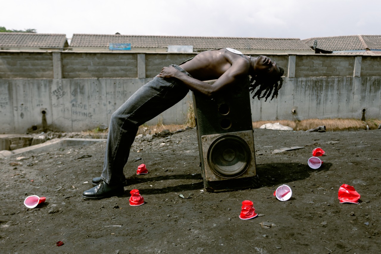 A young man leans back on a large speaker in a junkyard.