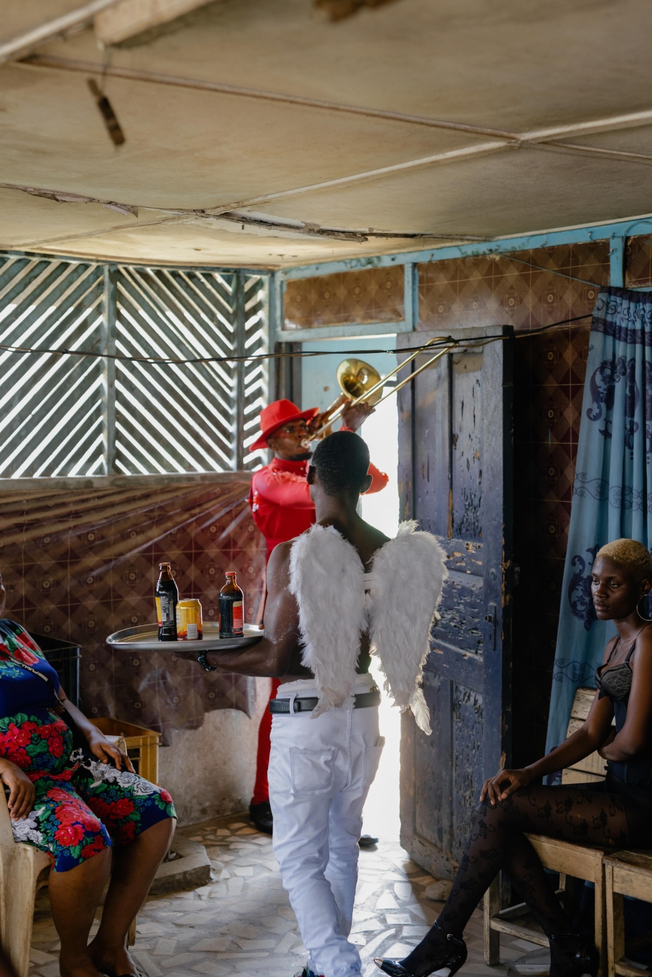 A man with white wings carries a tray of beverages inside a bar, while a man in red plays the trombone.
