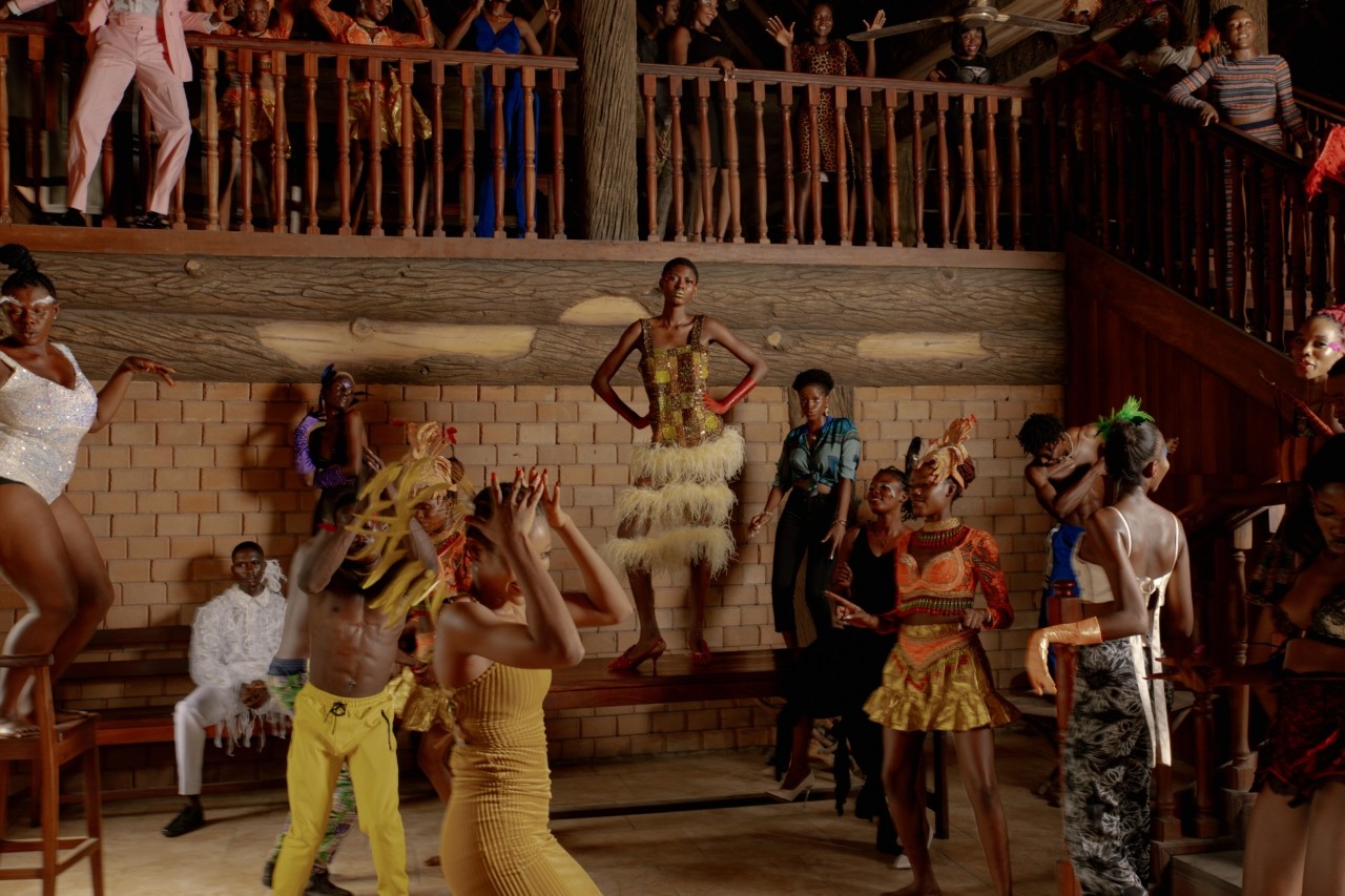 A carnival performer stands in the center of a bar with her hands on her hips while other performers dance around her.