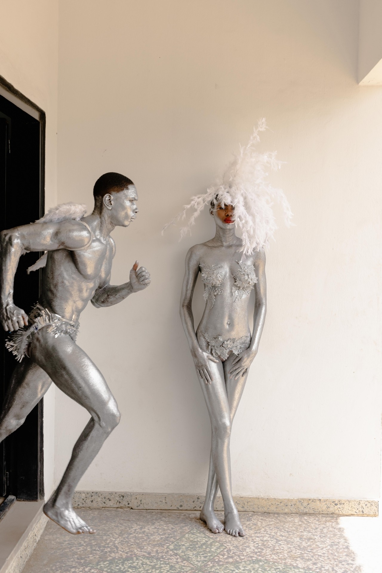 Two carnival performers are painted with silver body paint and adorned in white feathers. The male performer poses in a runners stance while the female performer leans against the white wall.