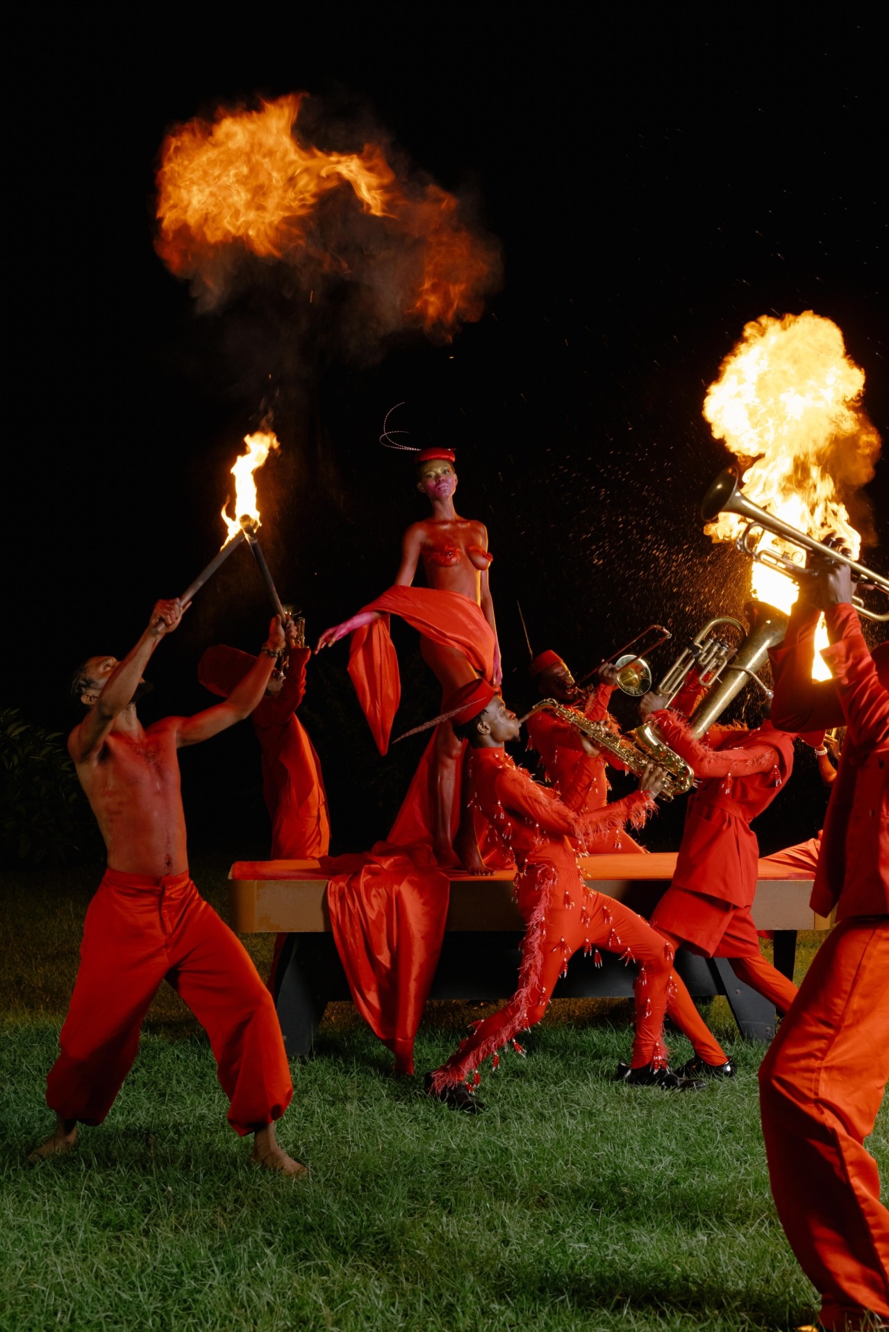 A female carnival performer is being carried on a flat piece of wood while the rest of the band plays instruments around her and holds firey sticks. They are all dressed in red.