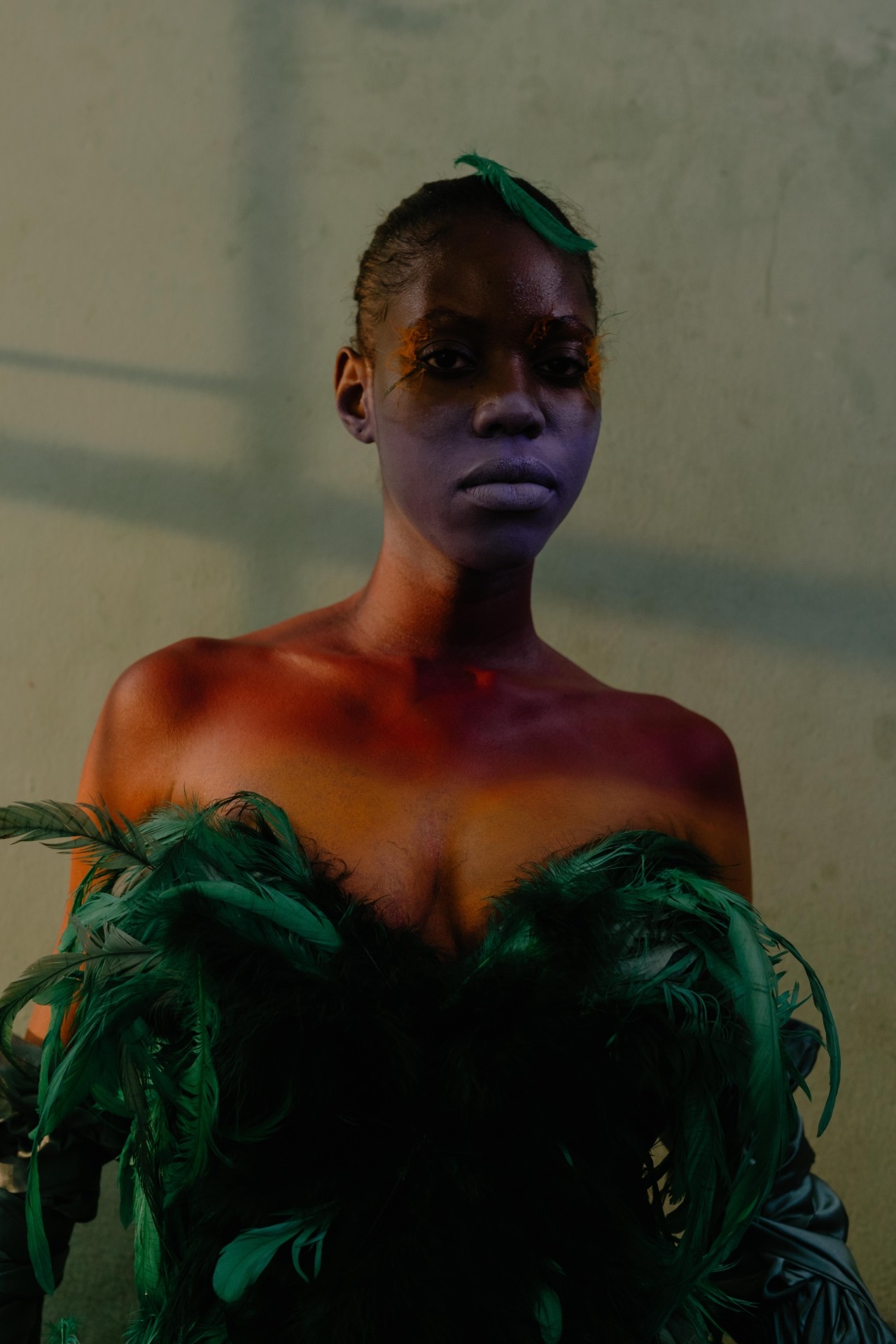 A woman with glittery face paint and a green feathery dress stands in a bedroom and looks into the camera.