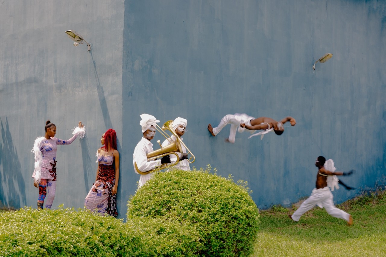 Six carnival performers convene outside a blue building. Two of them are having a conversation, two are holding instruments, and two are doing flips against the wall.